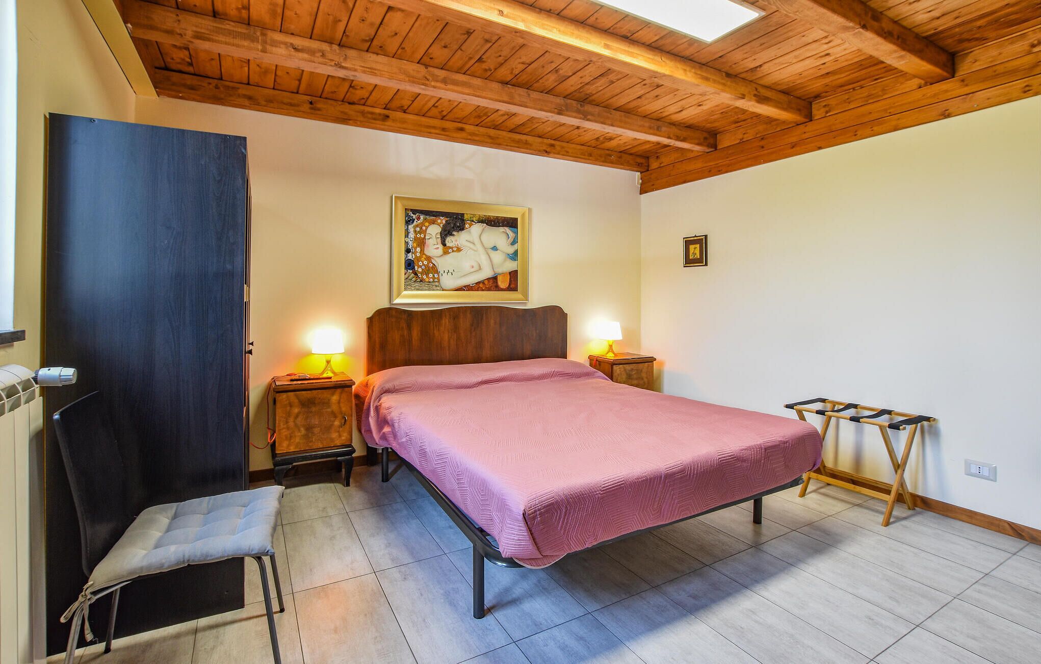 A bedroom with a bed, two bedside tables, a chair, a wooden bench, a painting on the wall, and a skylight.