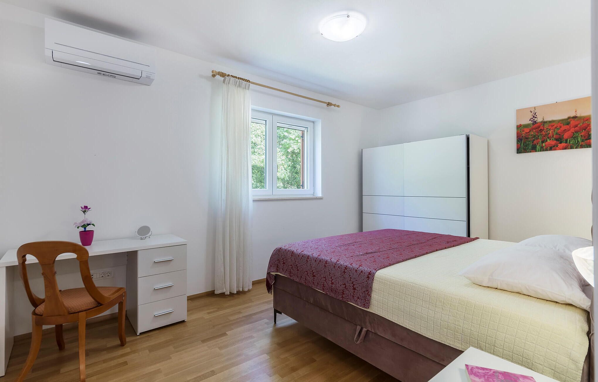 A bedroom with a bed, a desk with drawers, a chair, a window with curtains, and an air conditioning unit.