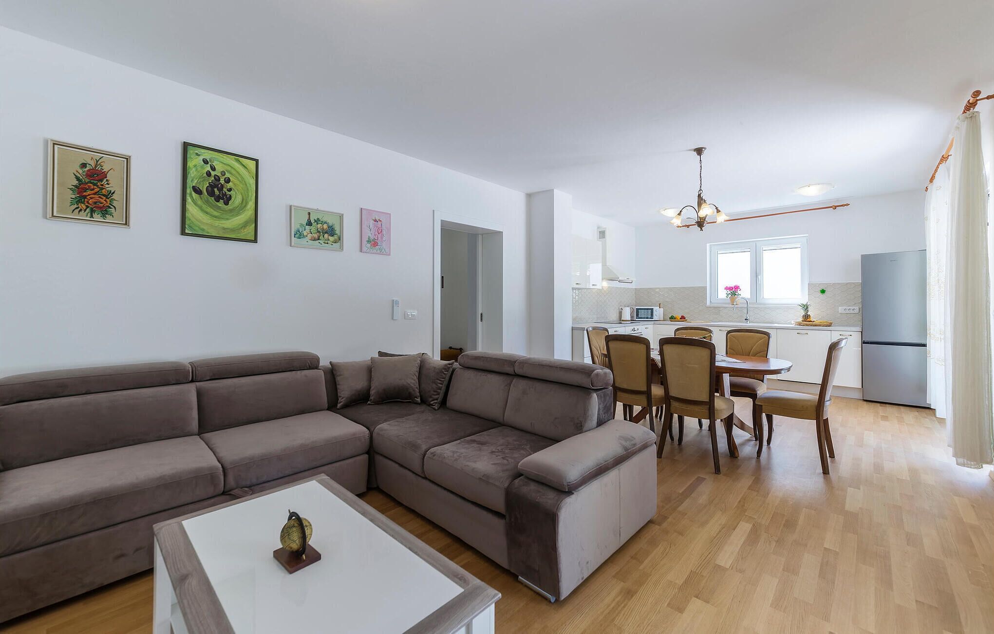 Un salon moderne avec un canapé d’angle, une table à manger avec des chaises, une cuisine en arrière-plan et des œuvres d’art accrochées aux murs.