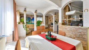 Un coin repas avec une table dressée pour deux personnes, un coin cuisine équipé d’une cuisinière et d’étagères, et un salon avec un canapé et une fresque murale.