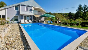 Une piscine entourée d’une maison et de verdure, avec des chaises longues et des parasols.