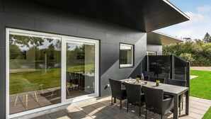 A modern outdoor patio with a dining table and chairs, a glass door leading to a garden, and a covered seating area.