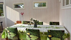 Ein Essbereich mit einem Tisch, bedeckt mit einer Tischdecke mit Blättermotiv, umgeben von Stühlen mit passenden Kissen. Zwei Fenster mit weißen Rahmen und eine kleine Vase mit Blumen an der Wand sind ebenfalls zu sehen.