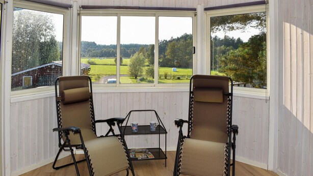 Ein Zimmer mit zwei Klappstühlen und einem kleinen Tisch, große Fenster mit Blick auf eine grüne Landschaft.