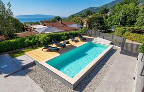 Une piscine rectangulaire dotée d’une terrasse en bois et de chaises longues, entourée d’une zone recouverte de gravier et d’une clôture en métal.