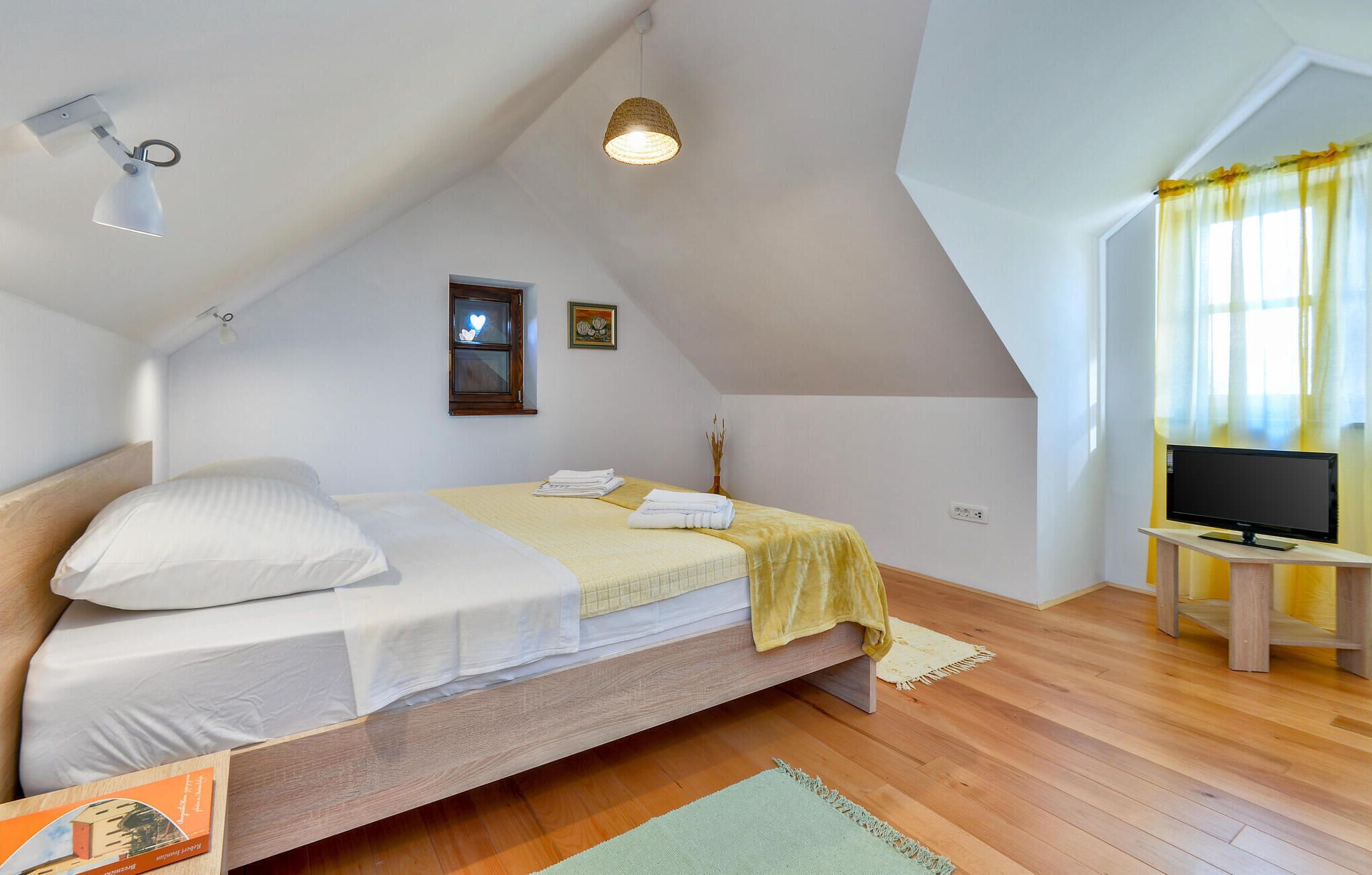 Un dormitorio con cama, televisor de pantalla plana, ventana con cortinas amarillas y lámpara en el techo.