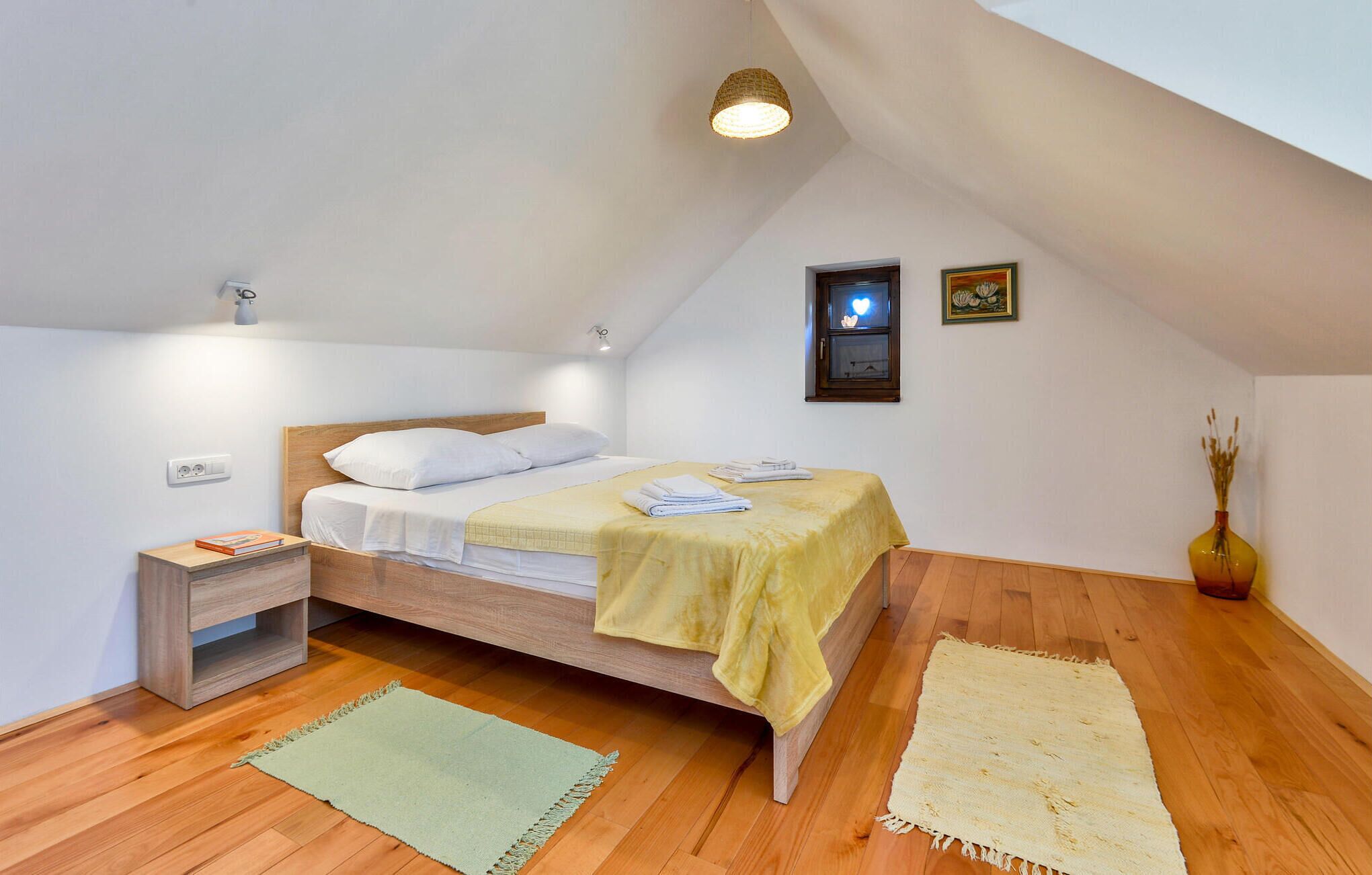 Un dormitorio con cama de madera, una mesita de noche, una lámpara de pared, un cuadro enmarcado y un jarrón con plantas secas.