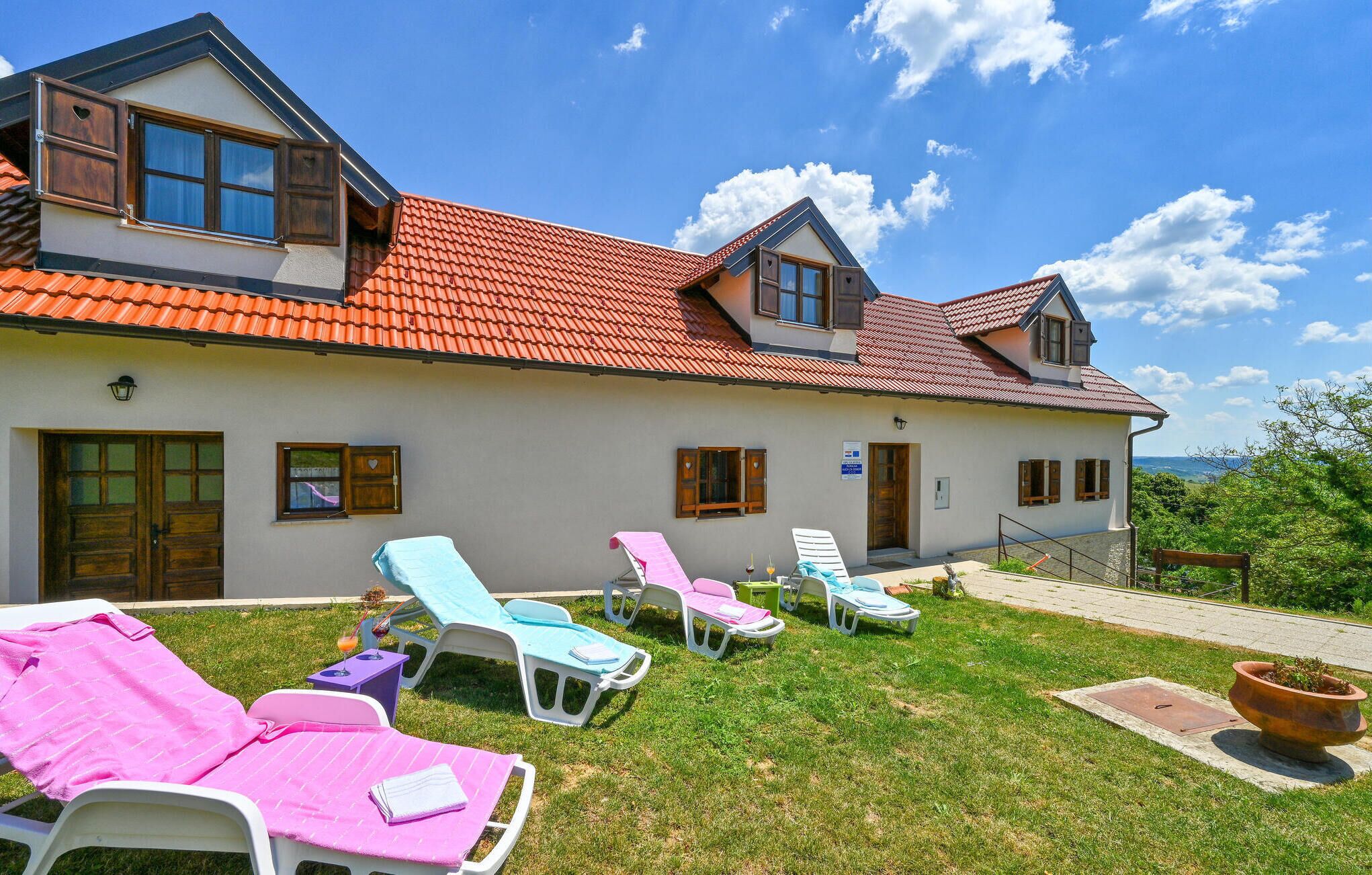 Een twee verdiepingen tellend huis met een rode pannendak, meerdere ramen en een gazon met enkele loungestoelen.