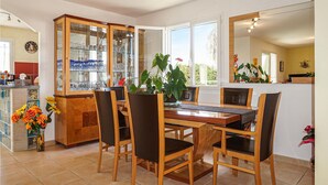 Un coin repas avec une table et des chaises en bois, un meuble vitré contenant de l’ustensile de cuisine, et une vue sur l’extérieur grâce à de grandes fenêtres.