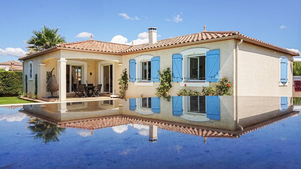 Une maison à deux étages, avec un toit recouvert de tuiles, des volets bleus et une piscine dans laquelle se reflète le bâtiment.