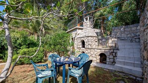 An outdoor patio with a stone-built oven, surrounded by lush greenery and a stone wall.