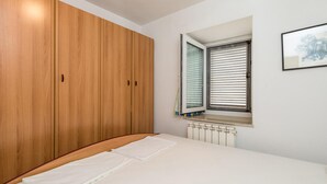 A bedroom with a wooden wardrobe, a bed with white bedding, a radiator, and a window with blinds.