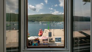 Vista a través de una ventana de un puerto deportivo con barcos, una colina verde y un quiosco con sombrillas.