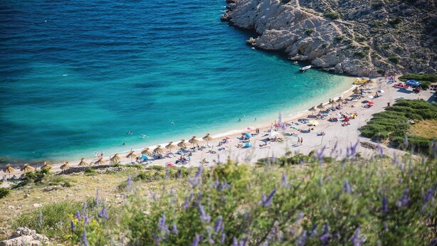 Una playa con aguas turquesas cristalinas, acantilados rocosos y una variedad de sombrillas y sillas de playa.
