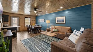 Un salon chaleureux avec un plafond en bois, un mur bleu, un canapé en cuir marron, une table basse et un coin repas avec une table et des chaises en bois.