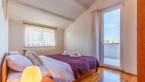 Quarto com cama, mesa de cabeceira iluminada por abajur, janela com cortinas estampadas e vista de edifícios através de uma porta de vidro.