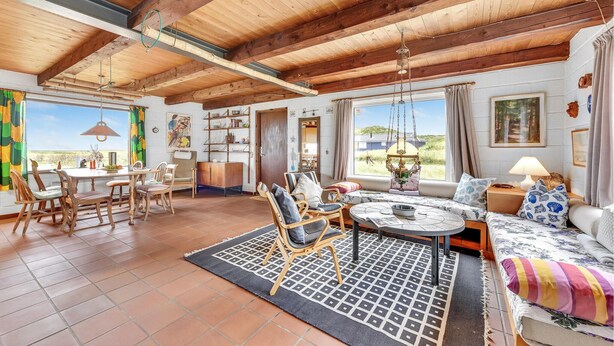 A spacious living area with a high wooden ceiling, a dining table with chairs, a sofa with cushions, and a large window offering a view of the outdoors.