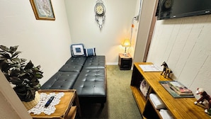 A compact living space with a sofa, a small table with a lamp, a TV mounted on the wall, and a decorative clock. - Deluxe Single Queen Suite Futon & Kitchenette (Salmon)