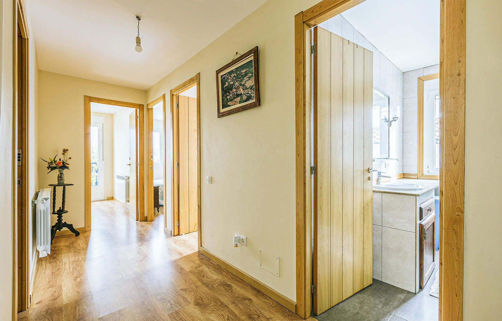 Un couloir avec un plancher en bois, un radiateur et un vase rempli de fleurs. Une porte donne accès à une salle de bain équipée d’un lavabo et d’un miroir.