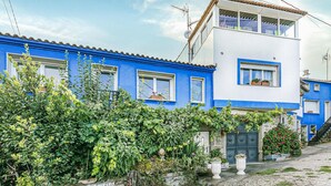 Un edificio de dos plantas con fachada azul y blanca, un muro de piedra con vegetación y un camino de grava que lleva a la entrada.