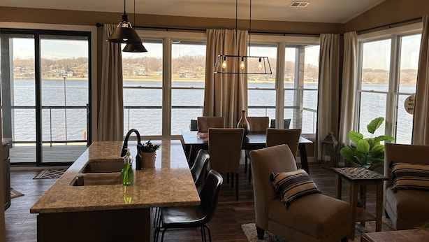 Een ruime woonkamer met een groot raam met uitzicht op een meer, een keukeneiland en een eethoek met stoelen en een tafel.