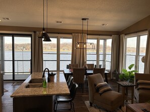 A spacious living area with a large window overlooking a lake, a kitchen island, and a dining area with chairs and a table. - Blue Suede Suites North (Port Byron)