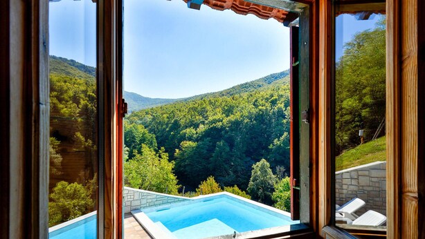 Vista desde una habitación con un ventanal grande que da a una piscina y una ladera boscosa.