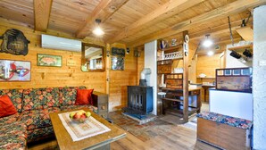 Interior acogedor de una cabaña de madera con sofá, una mesa con fruta, chimenea y estantes con varios objetos.