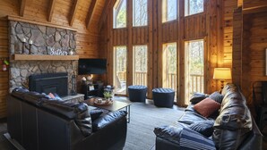 A cozy cabin interior with a stone fireplace, leather sofas, and large windows. - ❤️ of Hocking Hills 🌲 Hot Tub, Wifi, Close to Trails + Caves, 11 Secluded Acres (South Bloomingville)
