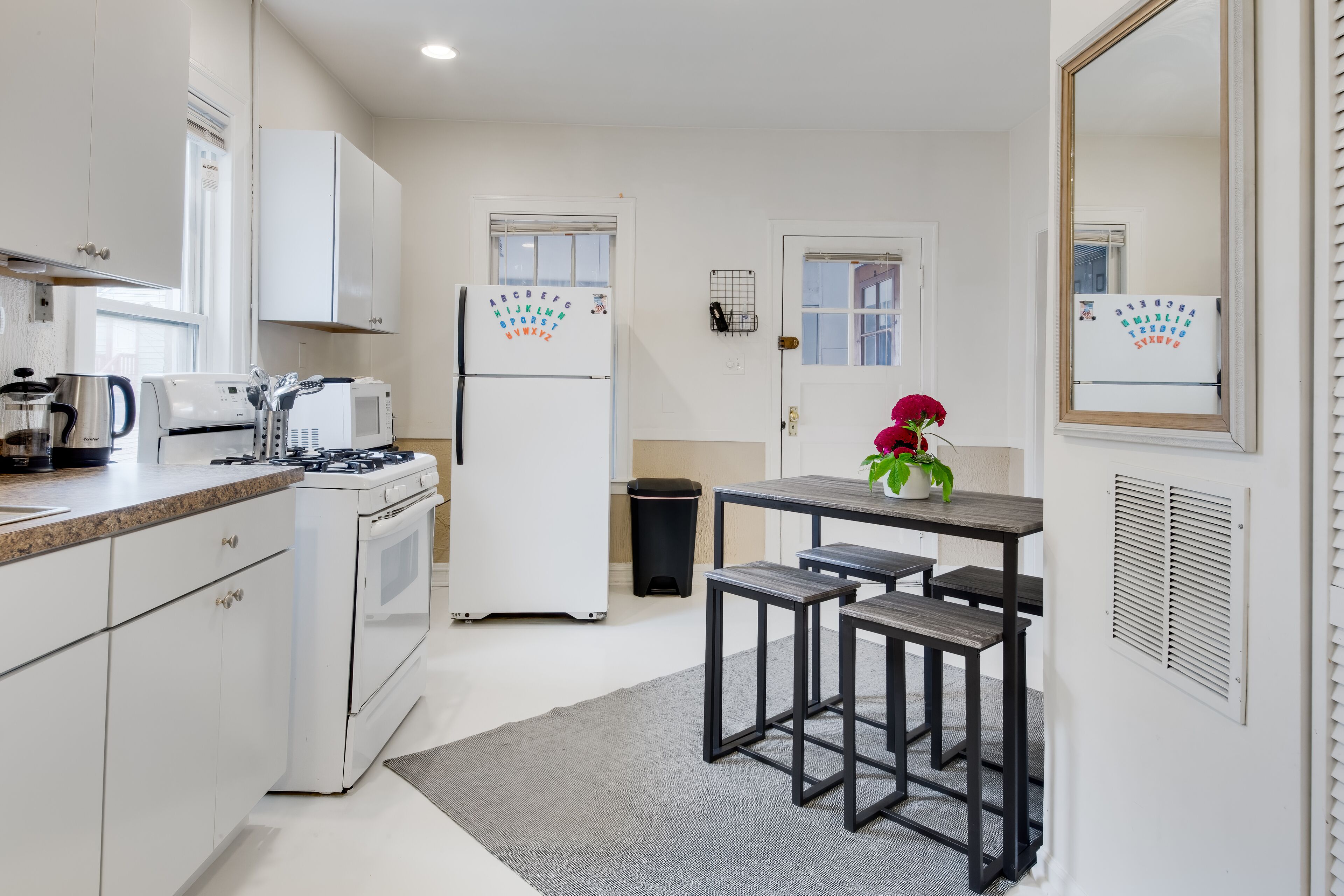 A modern kitchen with white cabinets, a gas stove, a microwave, and a refrigerator with children's alphabet magnets. There are two bar stools and a small table with a potted plant.