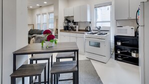 Une cuisine moderne avec des meubles blancs, une cuisinière à gaz, un four à micro-ondes et une étagère noire où sont rangées des casseroles. On y trouve également une table à manger avec des bancs et un vase de fleurs.