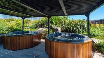 Deux bains à remous en bois, situés sous une terrasse couverte, offrent une vue sur une végétation luxuriante et un ciel bleu dégagé.