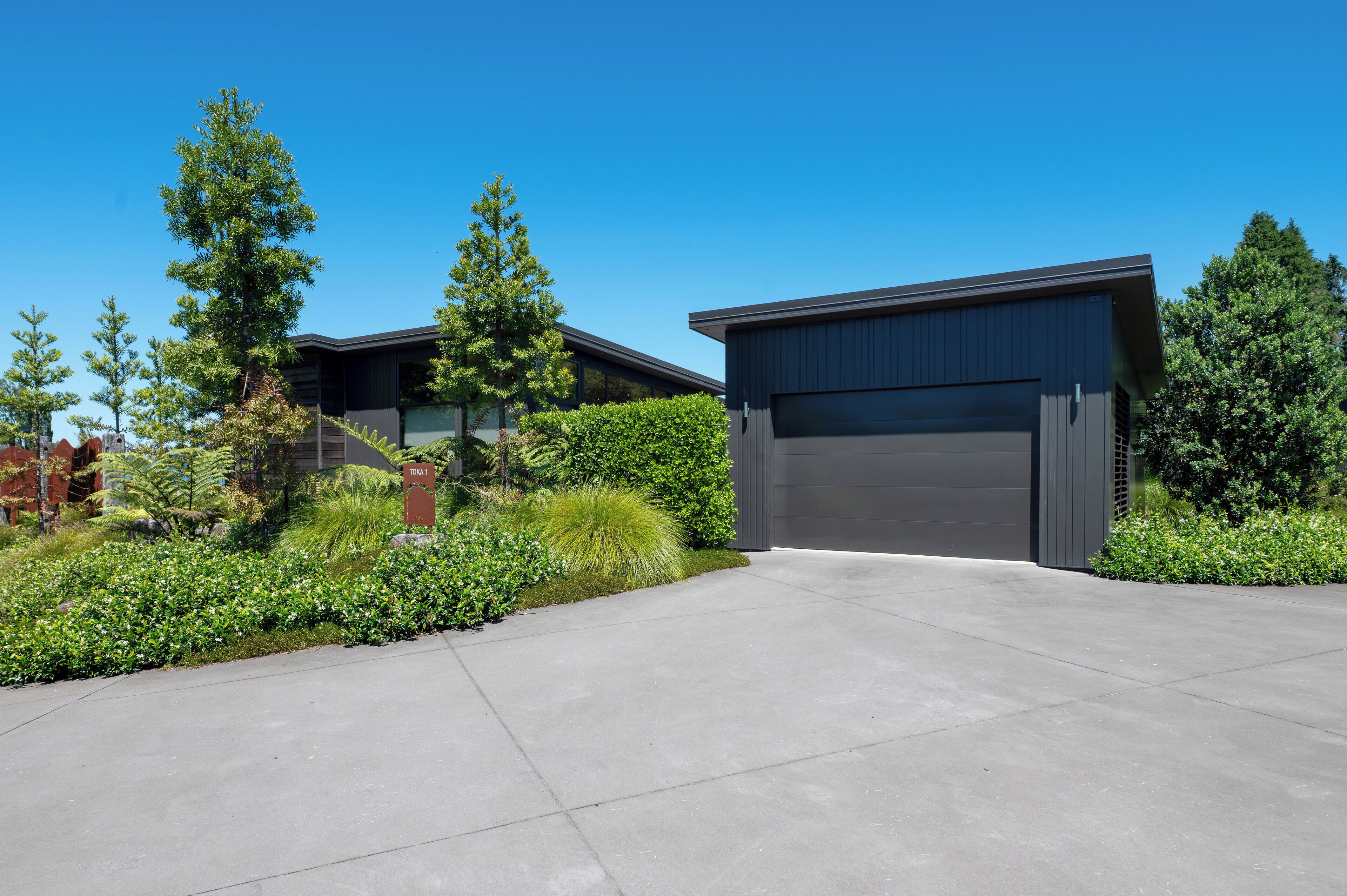 Une maison moderne à deux étages avec un garage, entourée d’un jardin bien entretenu et de grands arbres.