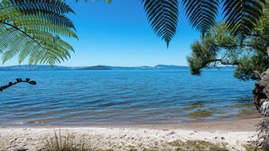 A beach with clear blue water, a sandy shore, and fern leaves framing the view. - Toka Ridge 1bd Lake View Lux Villa w/ HotTub (Hamurana)