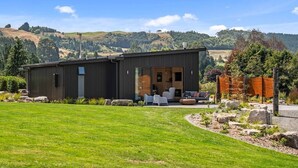 A modern building with a large patio area, a covered outdoor seating space, and a view of rolling hills in the background. - Toka Ridge 1bd Lake View Lux Villa w/ HotTub (Hamurana)