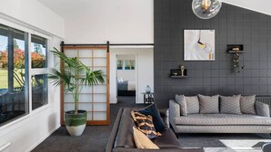 A modern living room with a dark grey wall, a grey sofa, a black leather sofa, a large potted plant, and a sliding door leading to another room. - Above Par Golf Course Villa with Hot Tub 4bd3.5bth (Rotorua)