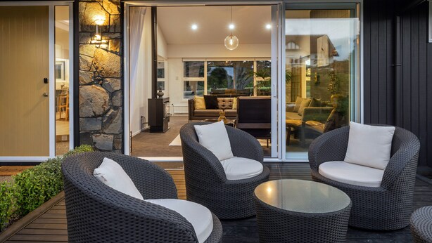 A modern outdoor seating area with wicker furniture and a glass table, leading to an interior space with a stone wall and a sofa. - Above Par Golf Course Villa with Hot Tub 4bd3.5bth (Rotorua)