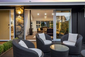 A modern outdoor seating area with wicker furniture and a glass table, leading to an interior space with a stone wall and a sofa. - Above Par Golf Course Villa with Hot Tub 4bd3.5bth (Rotorua)