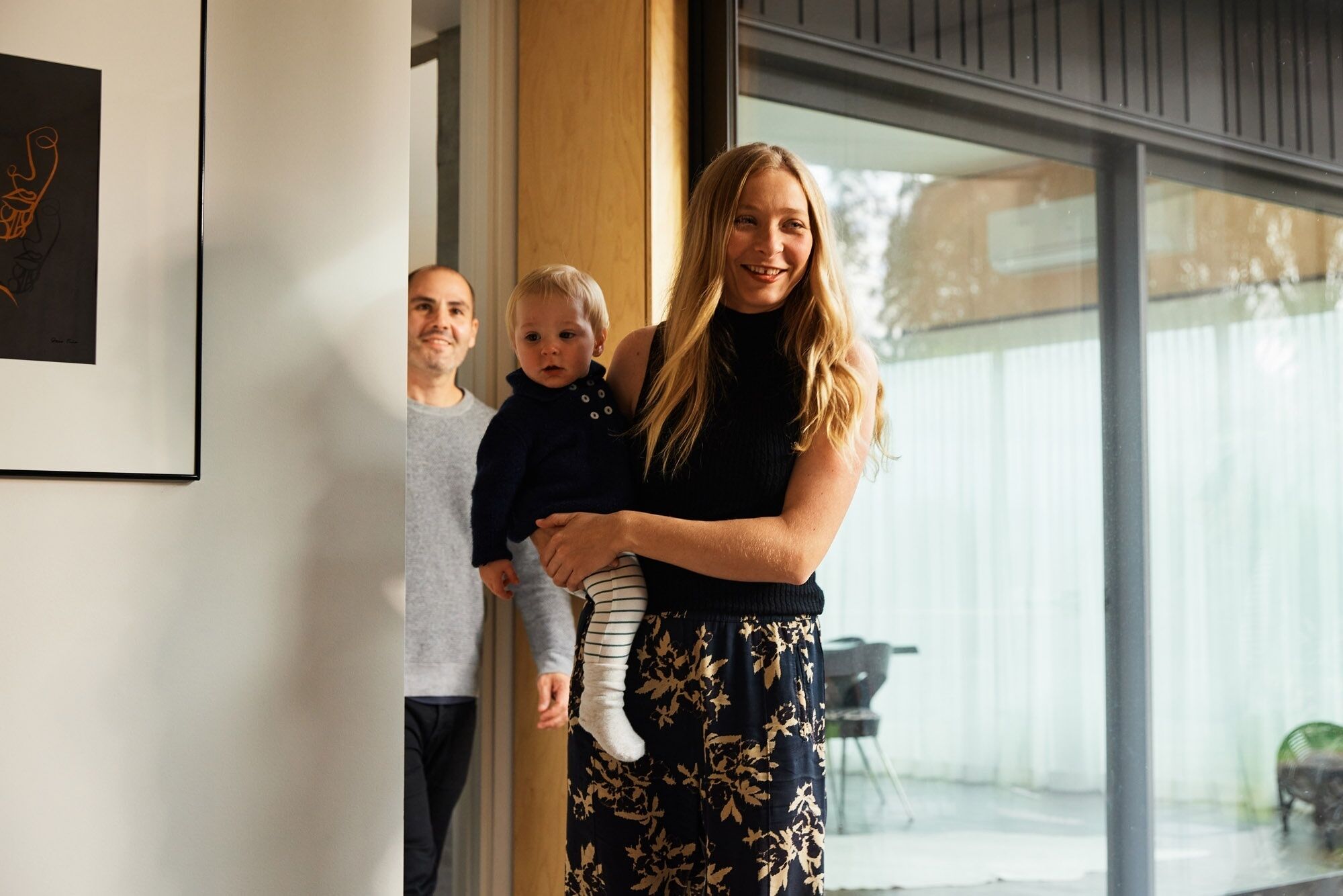 Une femme tenant un enfant, debout dans un encadrement de porte avec une grande fenêtre et un tableau encadré au mur.