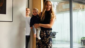 Une femme tenant un enfant, debout dans un encadrement de porte avec une grande fenêtre et un tableau encadré au mur.