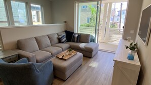 A modern living room with a sectional sofa, a small ottoman, and a coffee table. There is a window with blinds, a door leading outside, and a white countertop with a vase of flowers.