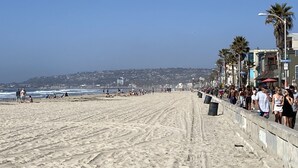 A beach with a concrete walkway, palm trees, and buildings in the background.