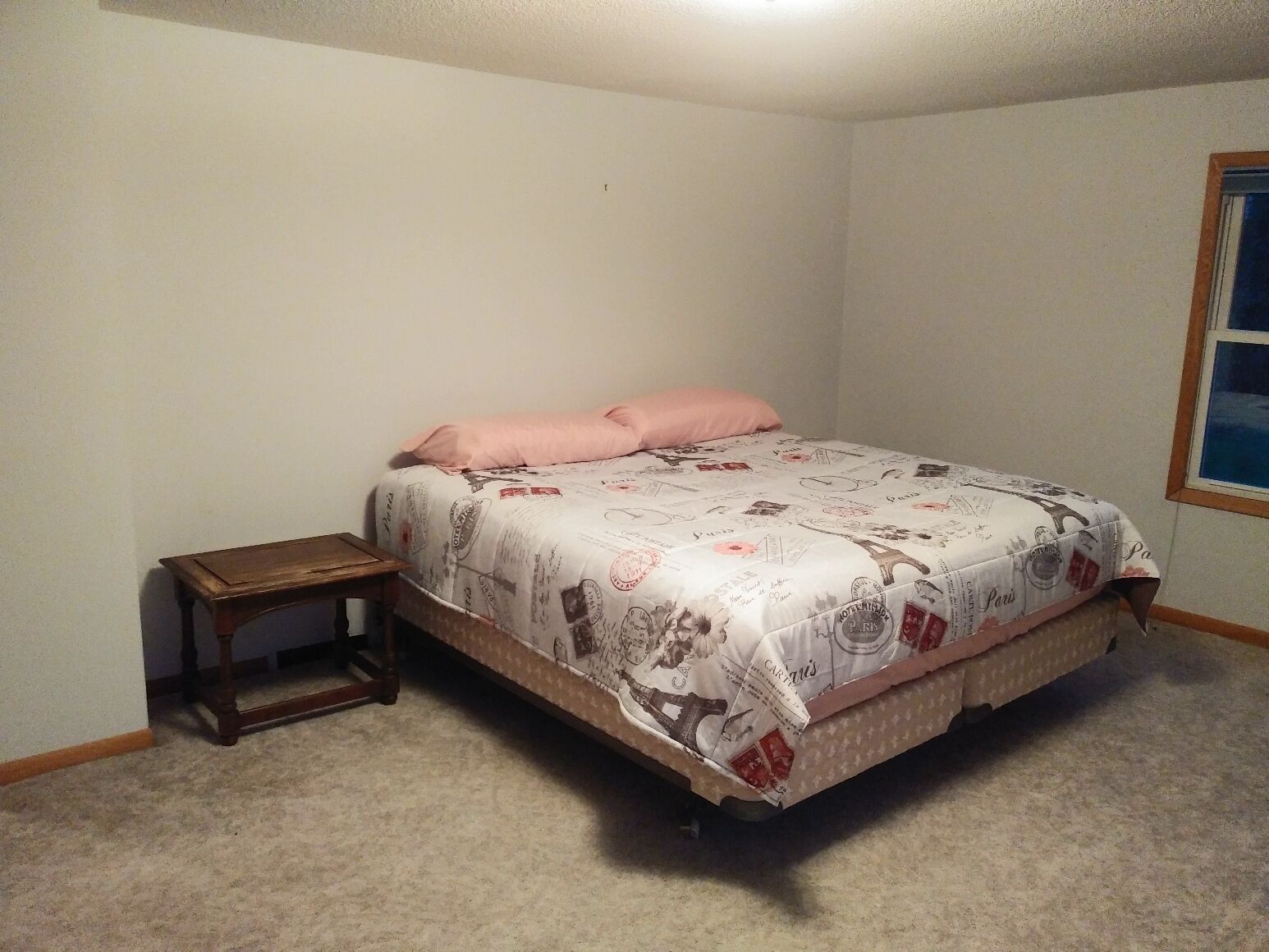A bedroom with a single bed, a small wooden nightstand, and a window with a view of the sky.
