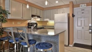 A kitchen with white cabinets, a granite countertop, a built-in oven, and a refrigerator. There are three bar stools with blue cushions.