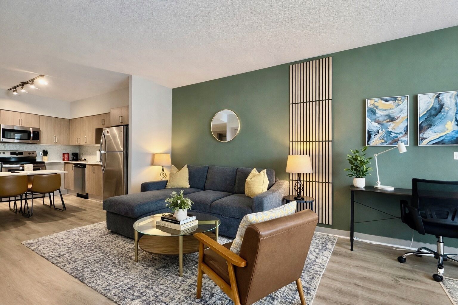 Un salon moderne avec un canapé gris, une table basse et un fauteuil marron. On y trouve également un coin repas avec une table et des chaises, et une cuisine à l’arrière-plan.