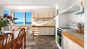 A compact kitchen with a view of the ocean, wooden cabinets, a stove, and a dining area with chairs. - Spindrift Lowerdeck BEACH FRONT location! Retro style 2 bed, Wifi, Nespresso++++ (Yamba)