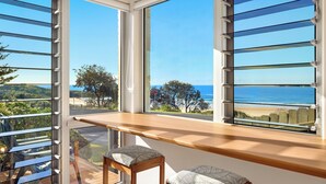 A room with large windows overlooking a beach and a hilly landscape. There is a wooden bench and two chairs with cushions. - Spindrift Lowerdeck BEACH FRONT location! Retro style 2 bed, Wifi, Nespresso++++ (Yamba)