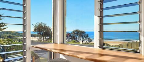 A room with large windows overlooking a beach and a hilly landscape. There is a wooden bench and two chairs with cushions.