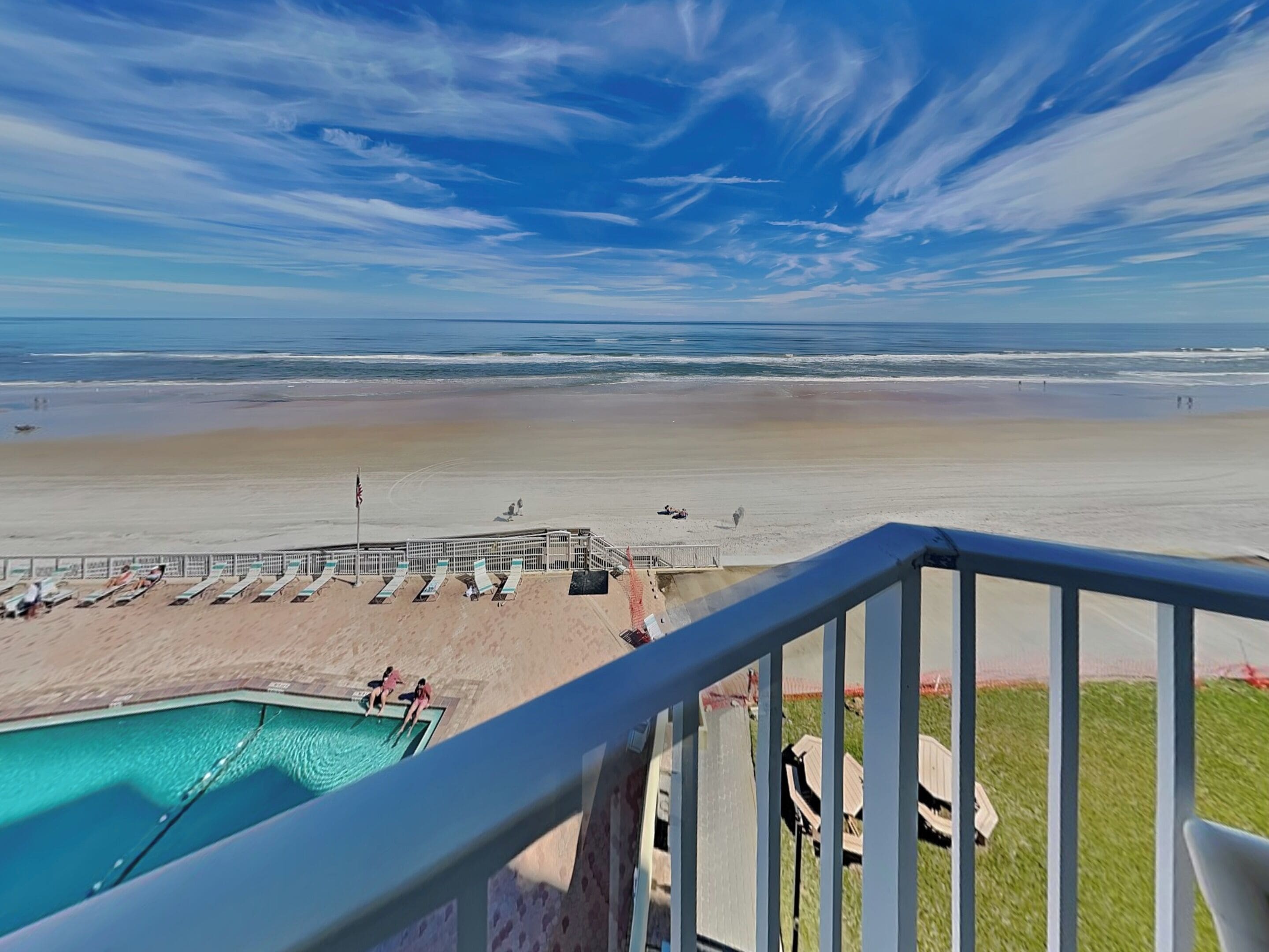 Blick auf einen Strand vom Balkon, mit Pool, Liegestühlen und einem Rettungsschwimmturm.