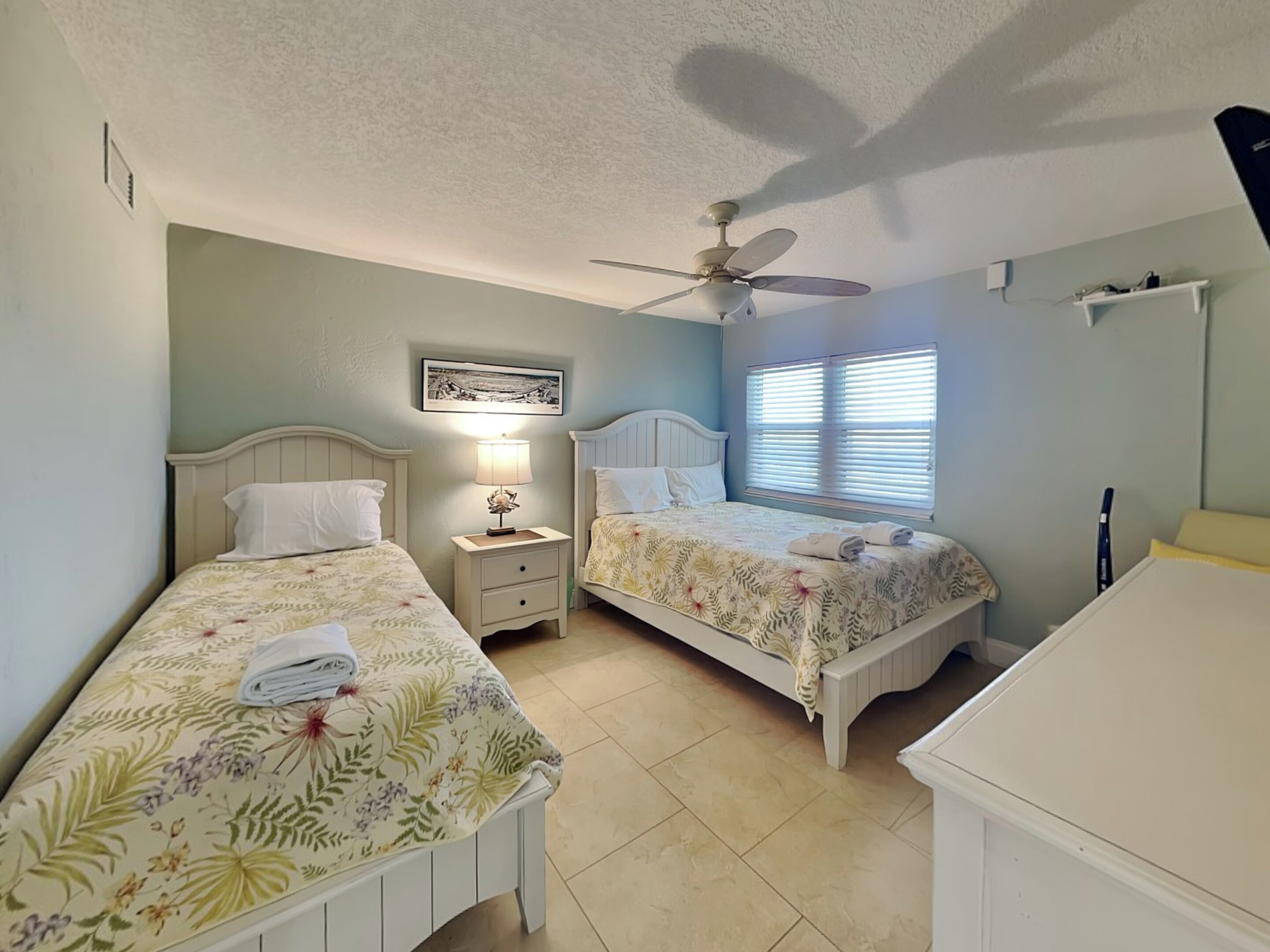 A bedroom with two beds, a ceiling fan, a window with blinds, and a wall-mounted picture.